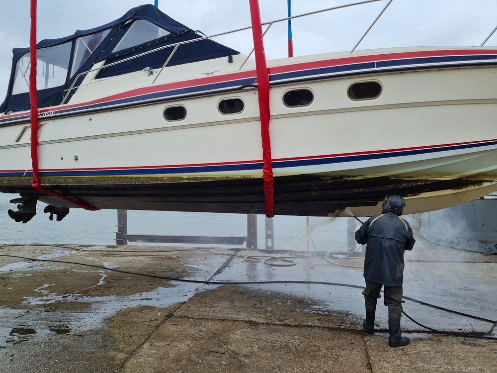 Motorboot in den Gurten eines Travellifts, ein Arbeiter reinigt das Unterwasserschiff mit dem Hochdruckreiniger